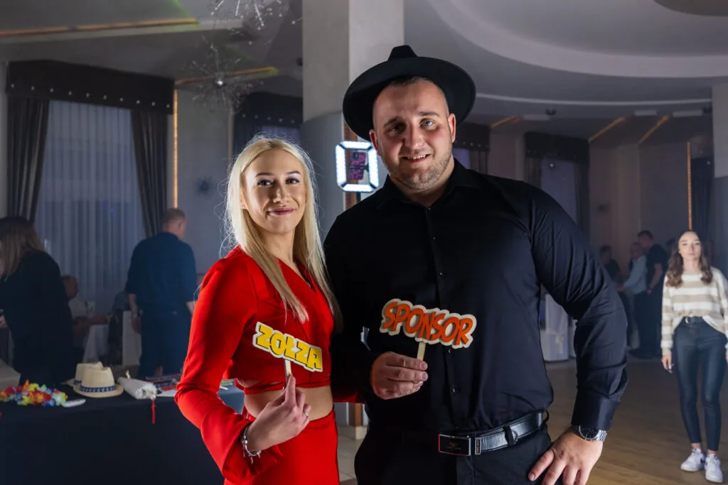 Blondynka w czerwonej marynarce i mężczyzna w czerni z drewnianym rekwizytem