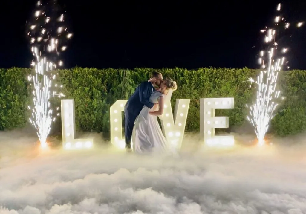 Para młoda całuje się przy napisie LOVE w ciężkim dymie, obok fontanny iskier